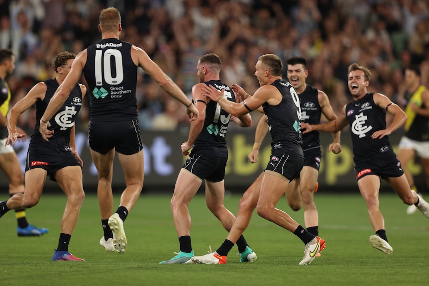 A group of AFL teammates grin widely as they rush in from all angles to get to their teammate who has just kicked a goal.