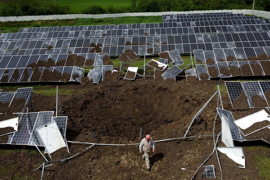 A middle-aged man walks over a large crater in the dirt amid solar panels on a solar farm.