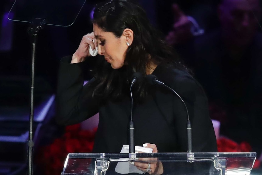 Dressed in black and standing at a podium, Vanessa Bryant turns her head away and holds a tissue to her eye.