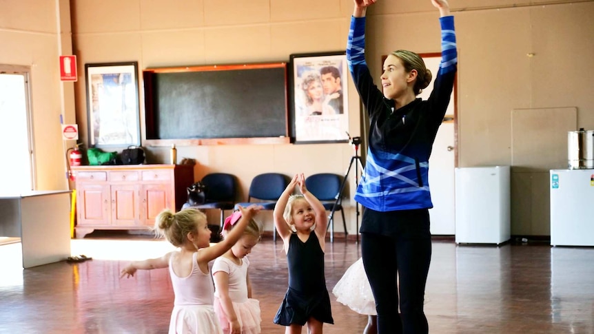 A young woman teaching four little girls dressed in ballet costumes how to twirl.
