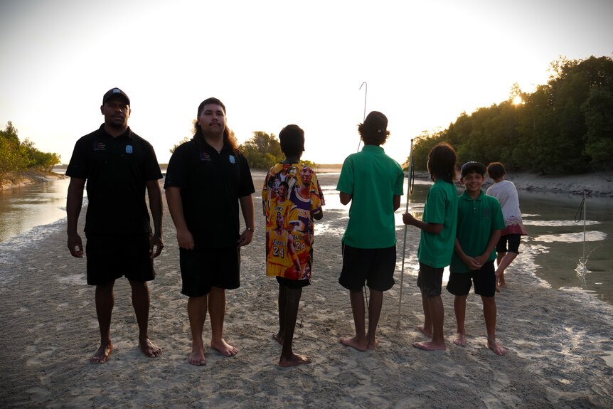 Two men facing forward and standing next to 5 boys who are facing away from the camera.