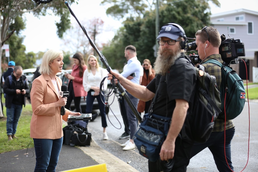 Woman holding a microphone talking to camera with a sound recordist nearby holding boom microphone.