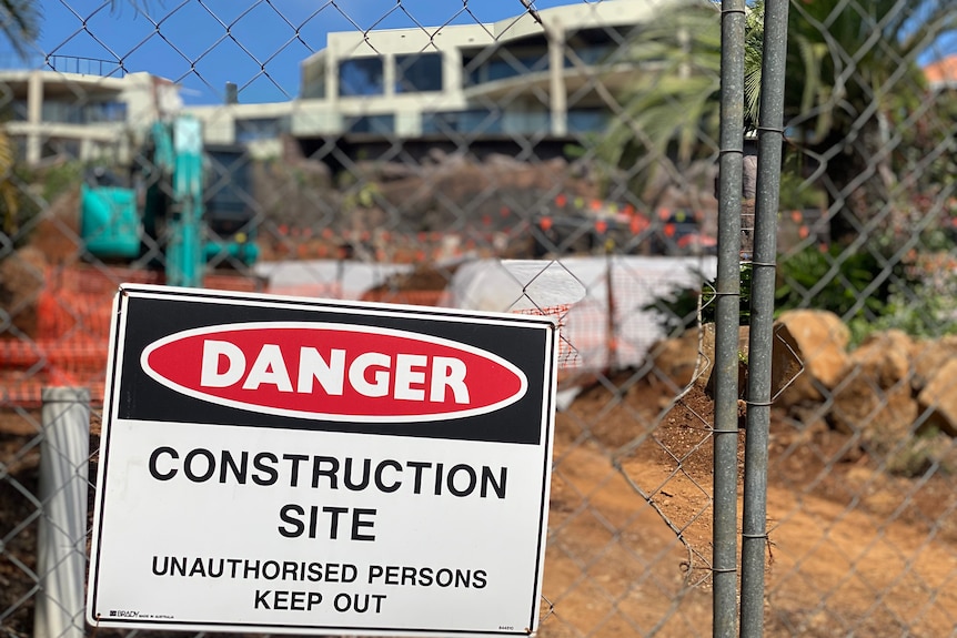 A construction site sign saying danger, a white building with glass visible behind the wire fence, blue sky.