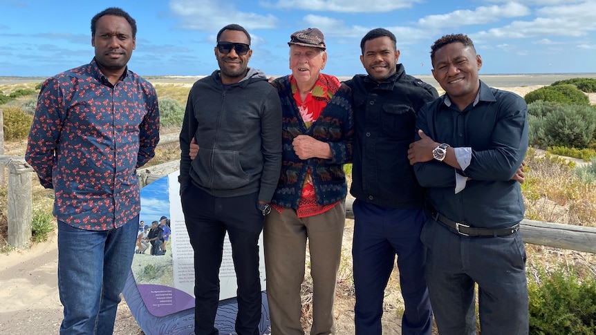 Four Vanuatu men and an old white man stand together smiling at a lookout point on a sunny day.