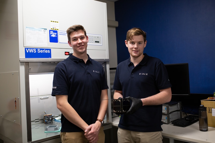 Two young men wearing dark polo shirts, one holding a small black cube.