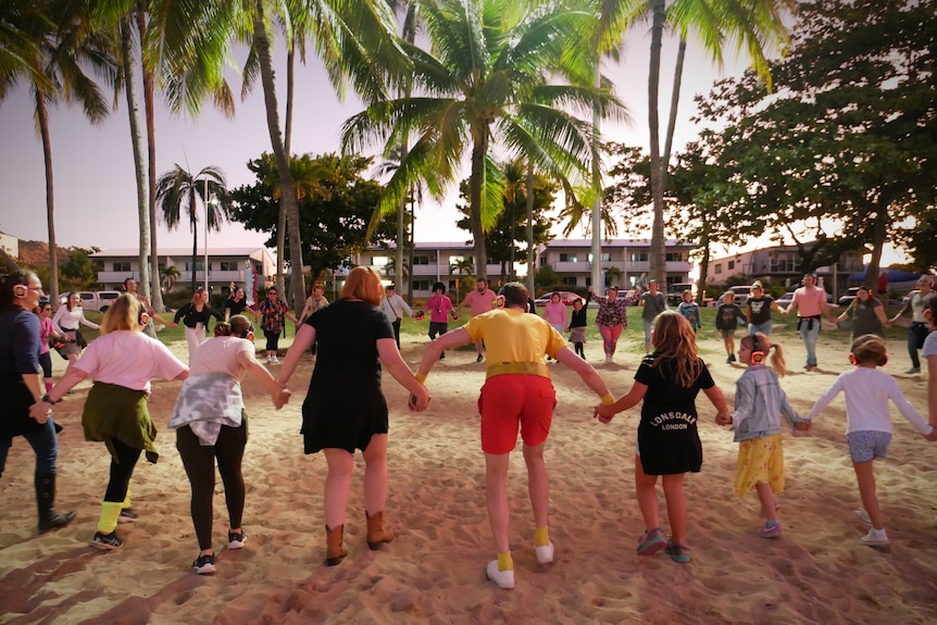 A group of people hold hands in a dance circle on a beach 