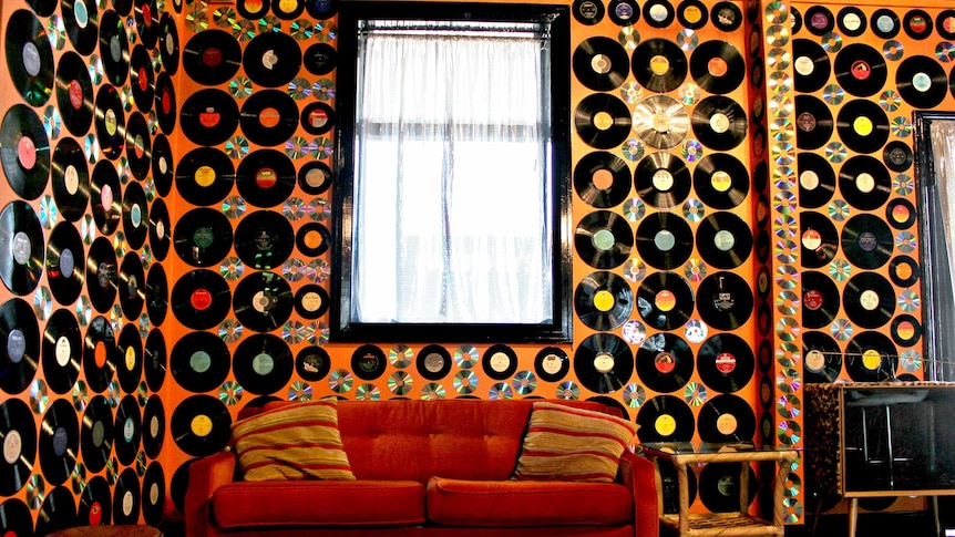 Bright orange wall covered in vinyl records with 1970's velvet couch in foreground.