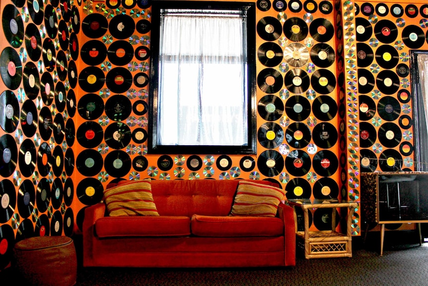 Bright orange wall covered in vinyl records with 1970's velvet couch in foreground.