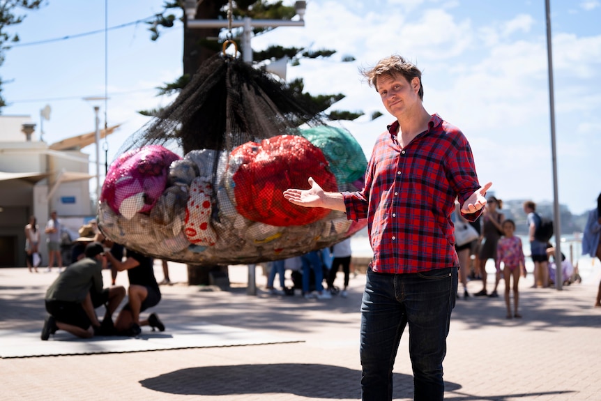 Craig Reucassel stands in front of 100kg of soft plastics at Manly Beach on a sunny day
