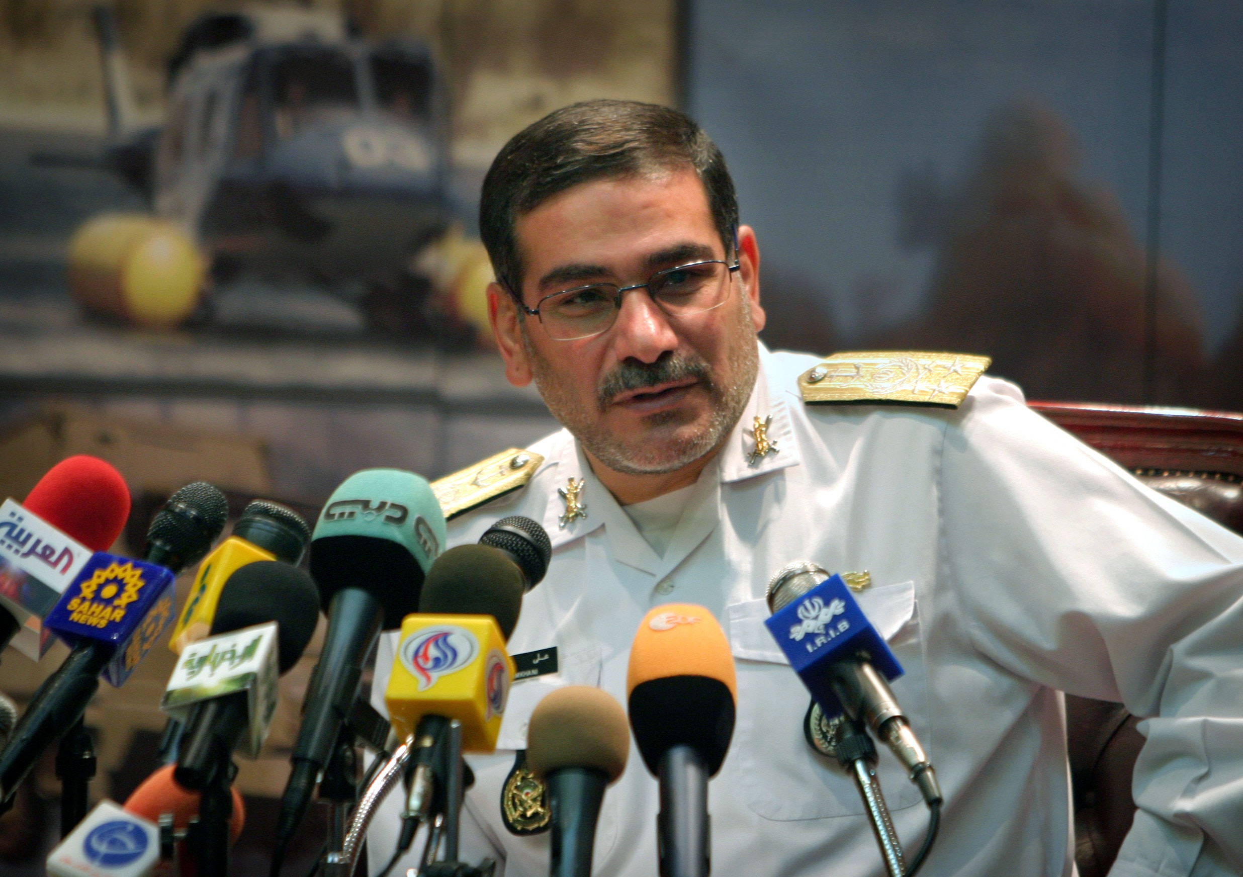 Ali Shamkhani  wearing a white military uniform leans toward a clutch of microphones set up on a desk.