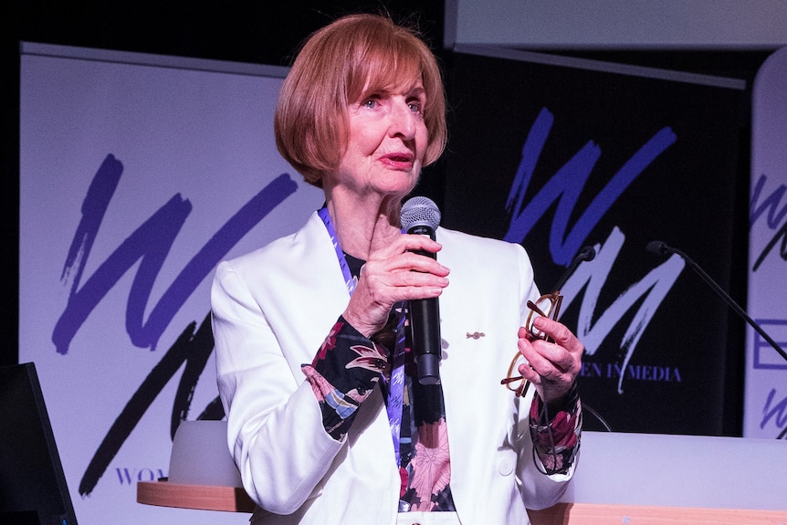 Caroline Jones wears a white pant suit speaking at a lecturn.