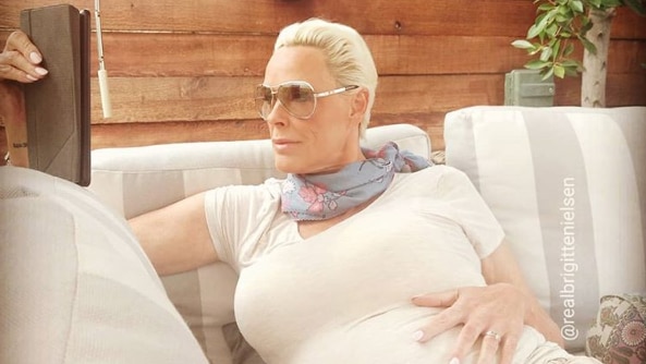 Brigitte Nielsen sitting on chair with one hand on pregnant stomach and the other holding a tablet.