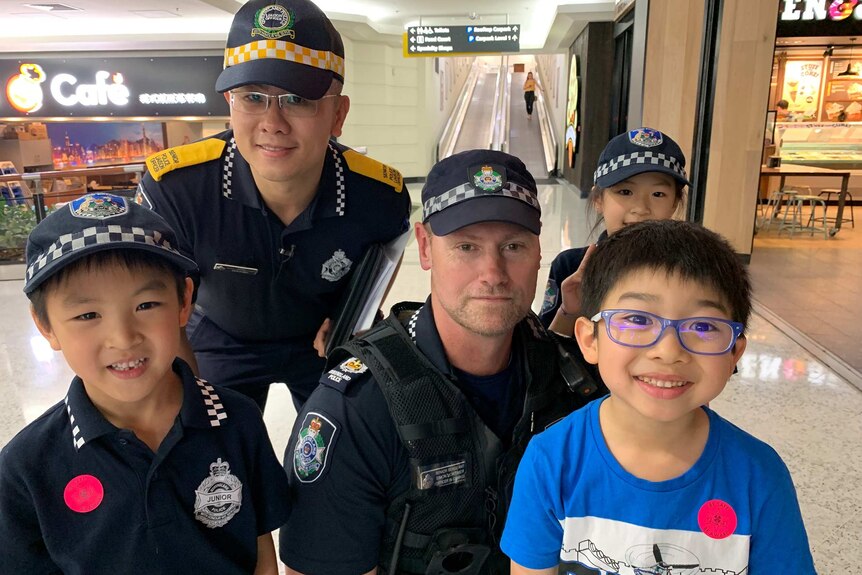 Two police officer stand with three children some of whom are wearing shirts and caps similar to those worn by police officers.