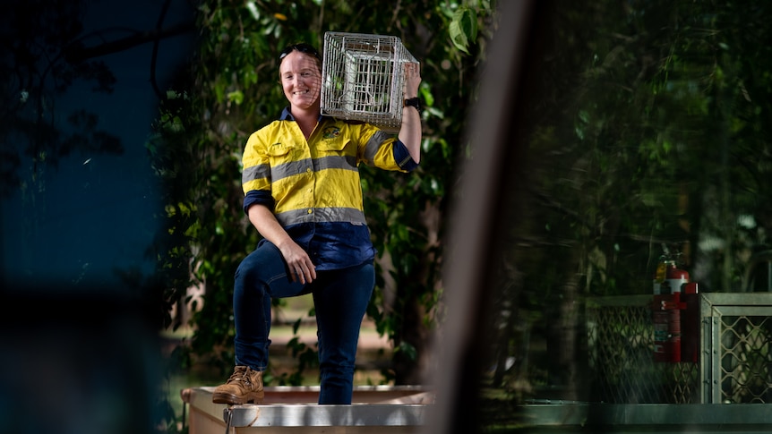 A woman in high vis smiling and holding an animal cage on one shoulder, with greenery in the background.