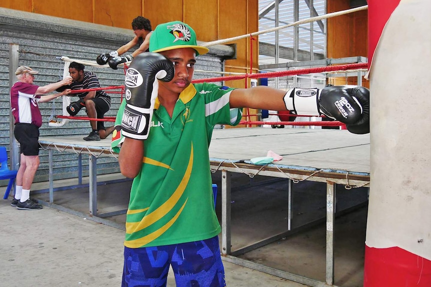 Indigenous boy punches a bag while int he background, coach gives another boxer a drink of water outside of the ring