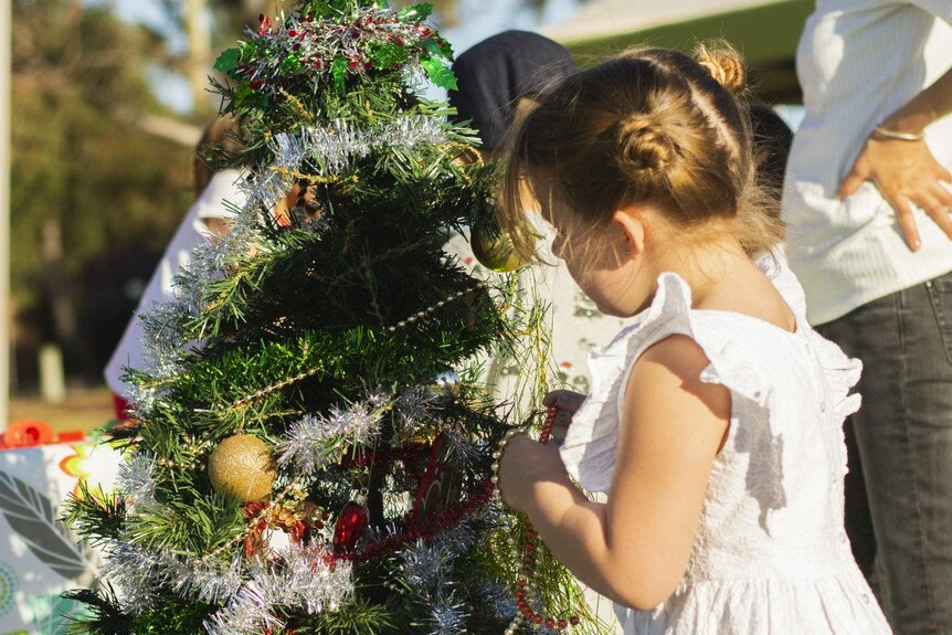 A young girl in a white dress looking putting tinsel on a small Christmas tree.