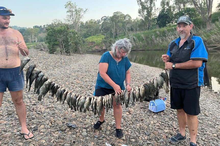 Two men hold up a long string filled with fish with a woman touching it in the middle.