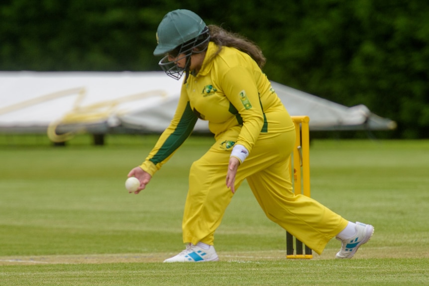 An Australian female cricketer is wearing a helmet and has the ball in her hand, preparing to bowl it underarm.