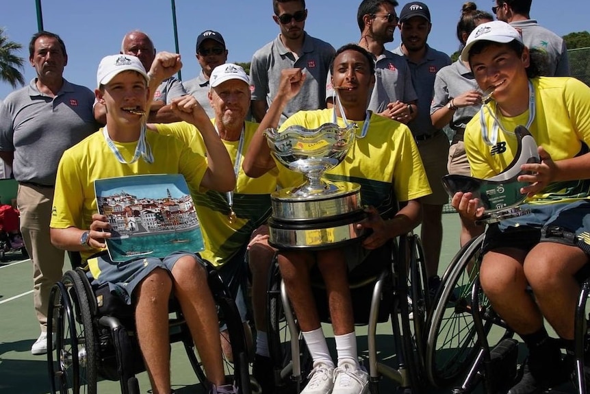 A trio of wheelchair tennis players smile as they hold medals and trophies after a tournament.
