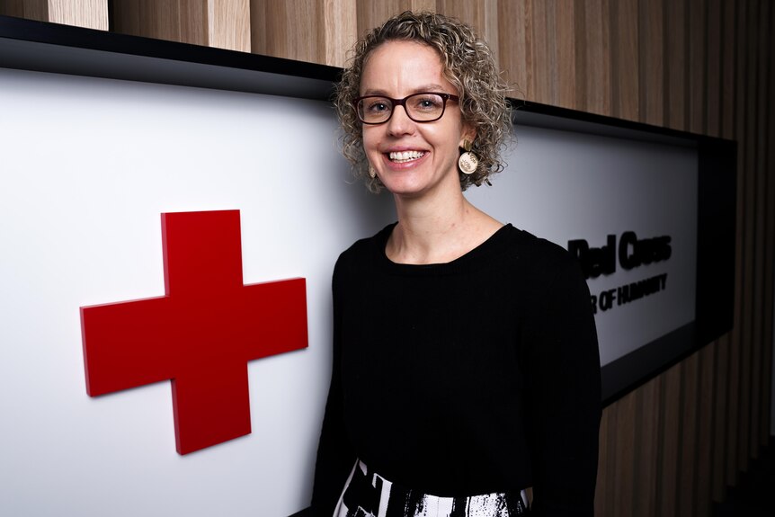 A woman wearing glasses smiles while standing next to a large red cross which is the Red Cross logo. 
