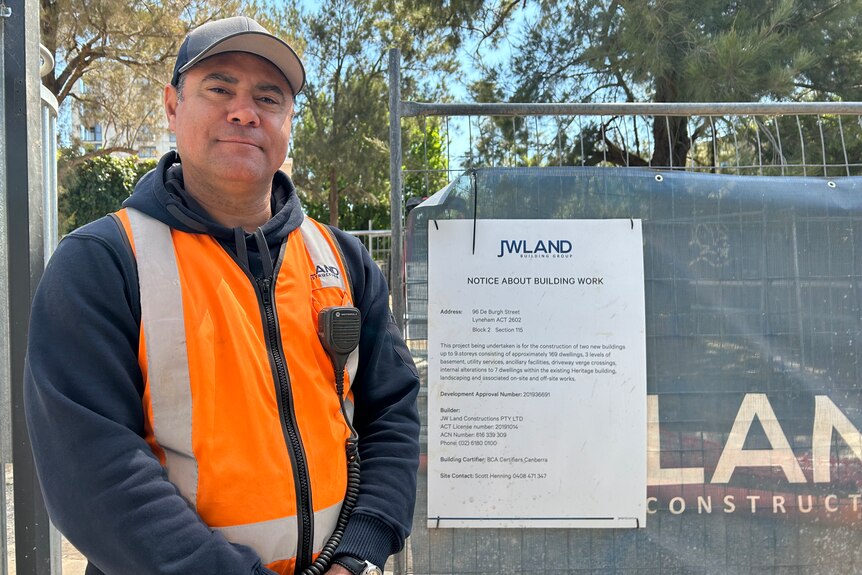 A man in a cap and a high-vis vest stands outside a construction site.