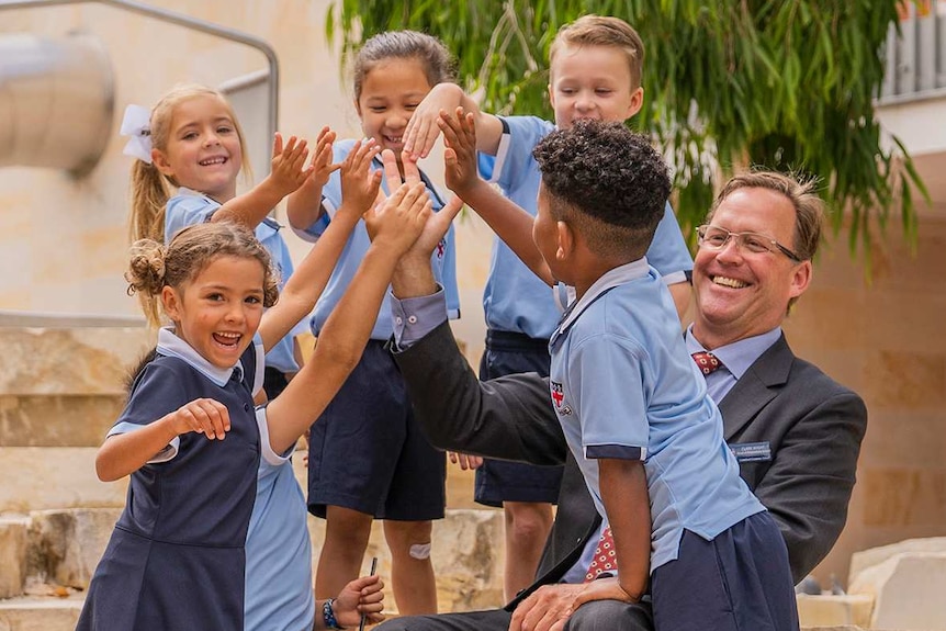 Guilford Grammar Preparatory School head Clark Wight sits in a playground smiling as a group of students high five each other.