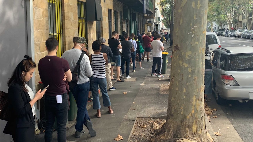 People stand in a line next to buildings on a footpath in Sydney