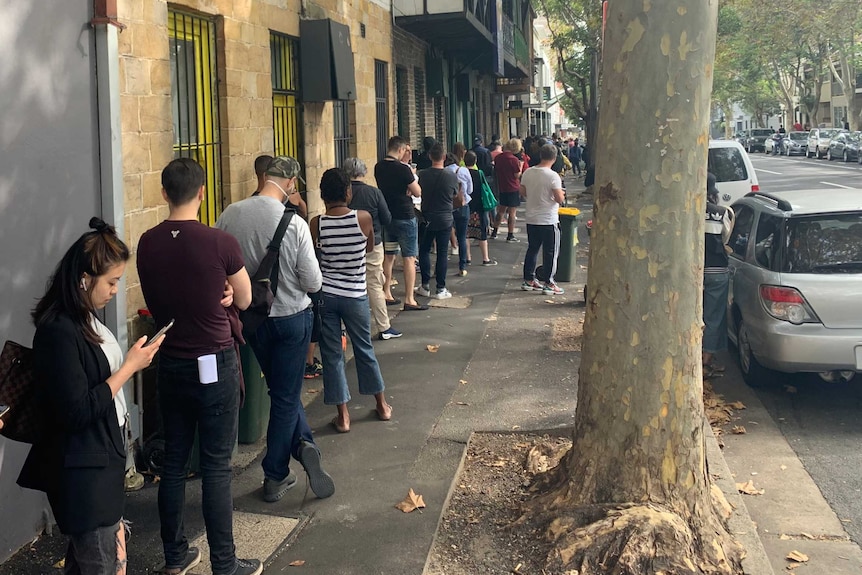 People stand in a line next to buildings on a footpath in Sydney