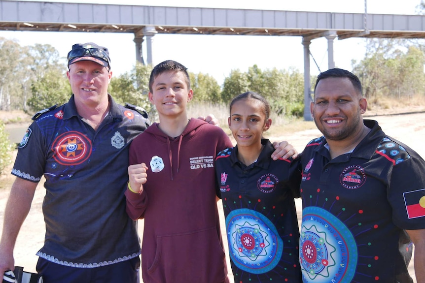 A policeman and boxing coach smile for a photo with two teenage boxers near a bridge in a park in Bundaberg North, Queenland.