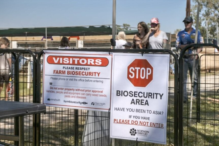 Two biosecurity related signs on a fence with people walking past