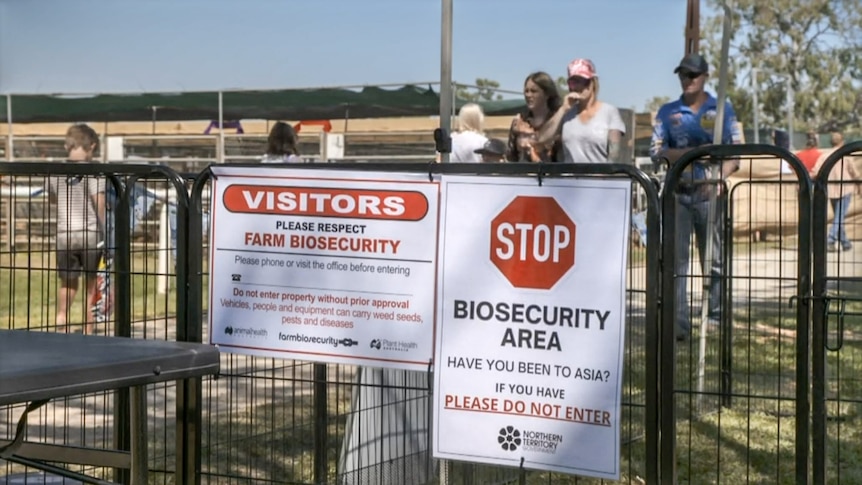 Two biosecurity related signs on a fence with people walking past