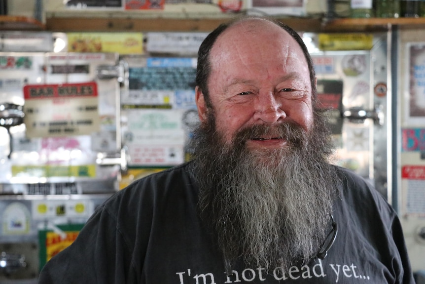 A man stands behind a bar in a black shirt and a grey beard.