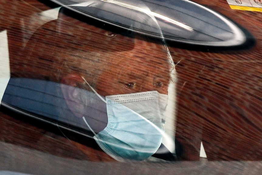 An elderly man wearing a face mask peers through a car window from the passenger's seat.