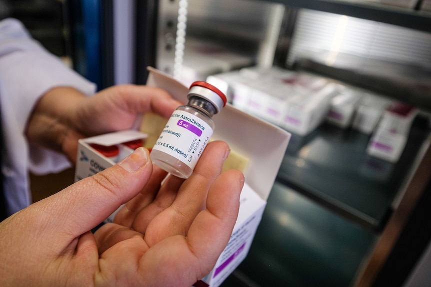 A hand holding a vaccine vial. AstraZeneca 5ml is printed on the label