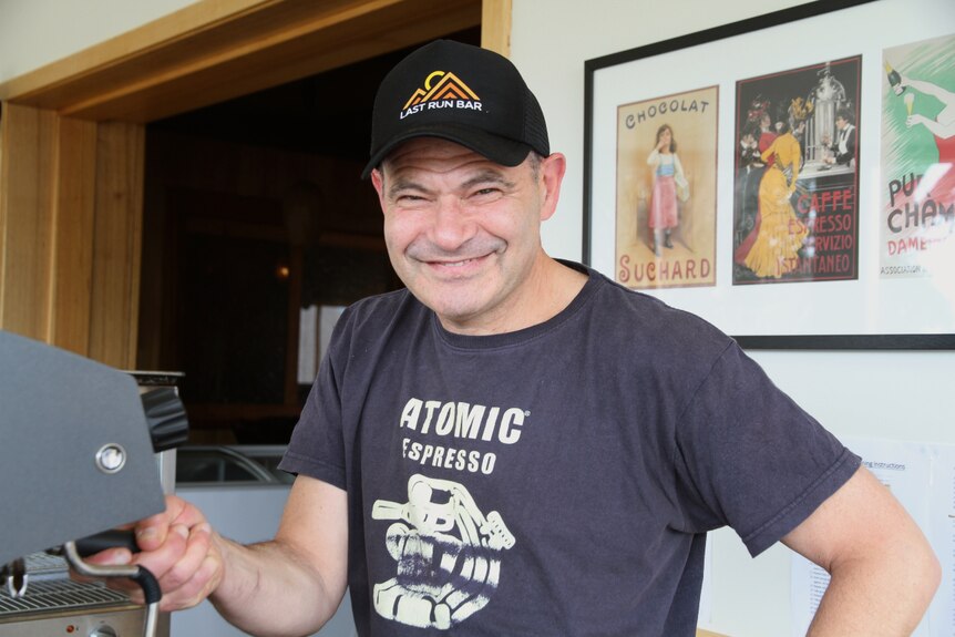 A man wearing a black cap smiles while he operates an espresso machine.