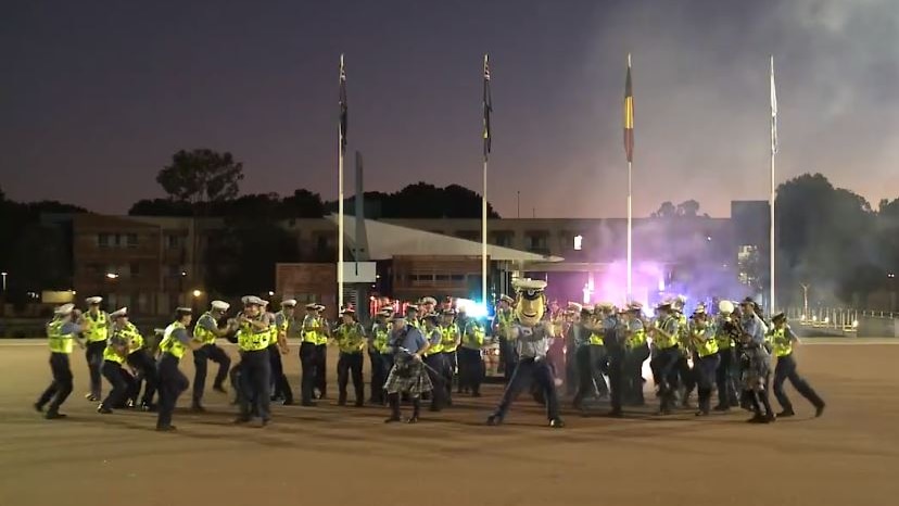 A group of police officers in hi-vis clothing dances in a courtyard at night with police sirens behind.