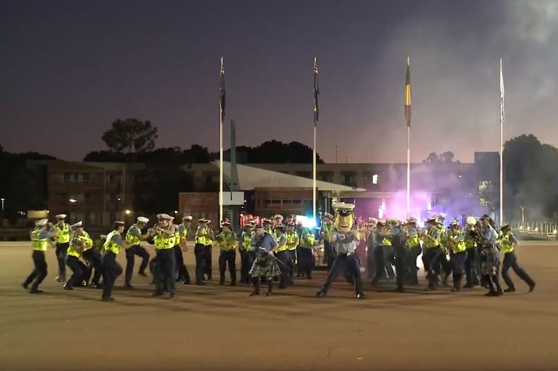 A group of police officers in hi-vis clothing dances in a courtyard at night with police sirens behind.