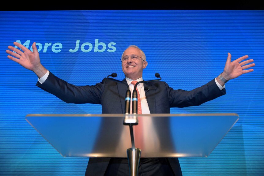 Malcolm Turnbull smiles and holds his hands wide open while standing at a podium.