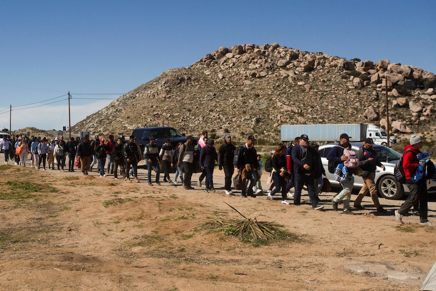 A queue of people walks along a road in front of a rocky hill.