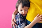Young boy with dark hair and a blue check shirt hugs the hip of a woman wearing a yellow jumper and blue jeans