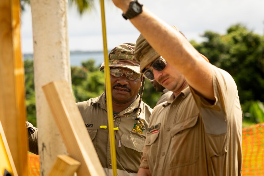 Two defence personnel measure a wooden beam on a construction sight. 