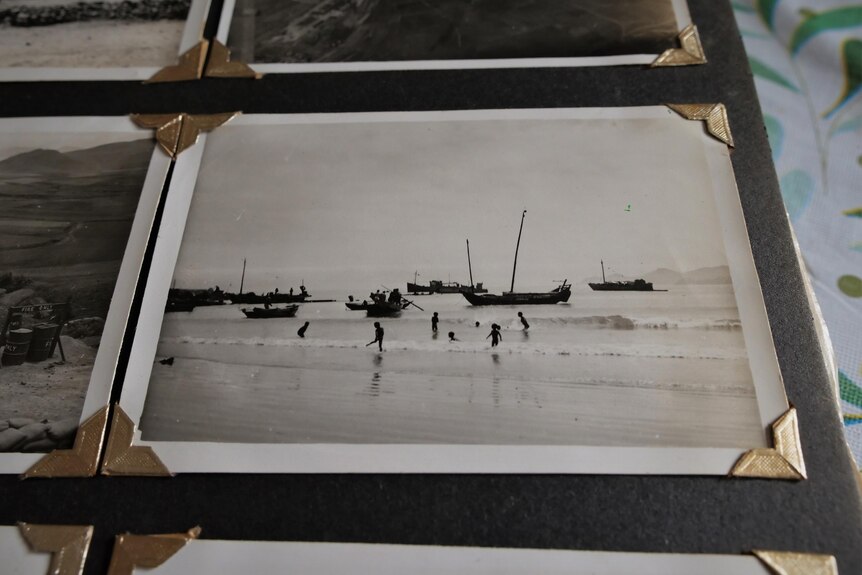 A black and white photo showing black silhouettes of children playing in the sea with boats in the background
