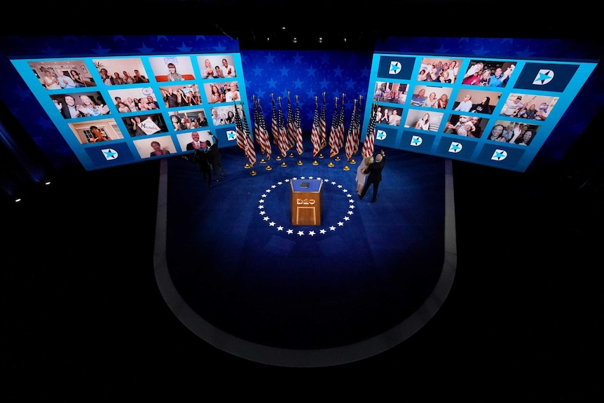 A man and woman stand on a stage with american flags looking up at two screens with multiple people in squares