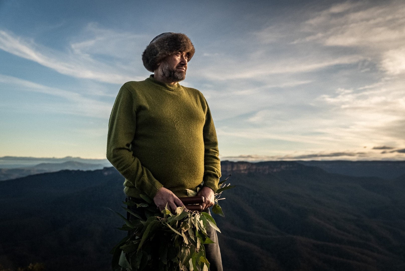 Man stands on a cliff top with sandstone cliffs and trees behind him, wearing a fur hat and holding a branch of gum leaves.