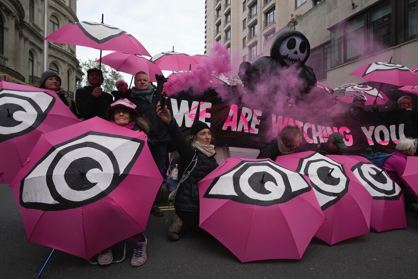 Environmental activists gather with pink umbrellas with eyeballs painted on them holding a sign that reads "we are watching you"