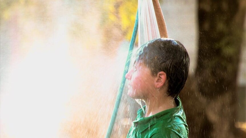 A young boy holds a hose above his head, while fully clothed.