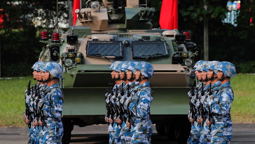 Soldiers in blue and white camo helmets and fatigues march past a tank and Chinese flags, lined up perfectly, and holding guns.