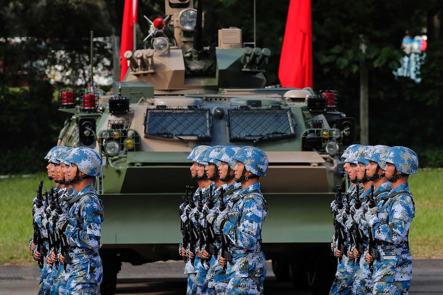 Soldiers in blue and white camo helmets and fatigues march past a tank and Chinese flags, lined up perfectly, and holding guns.