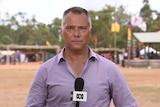 Stan Grant stands with an microphone in hand, staring down the barrel of the camera. There are Indigenous flags behind him