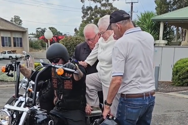 An old lady getting on the back of a motorcycle with two men helping her.